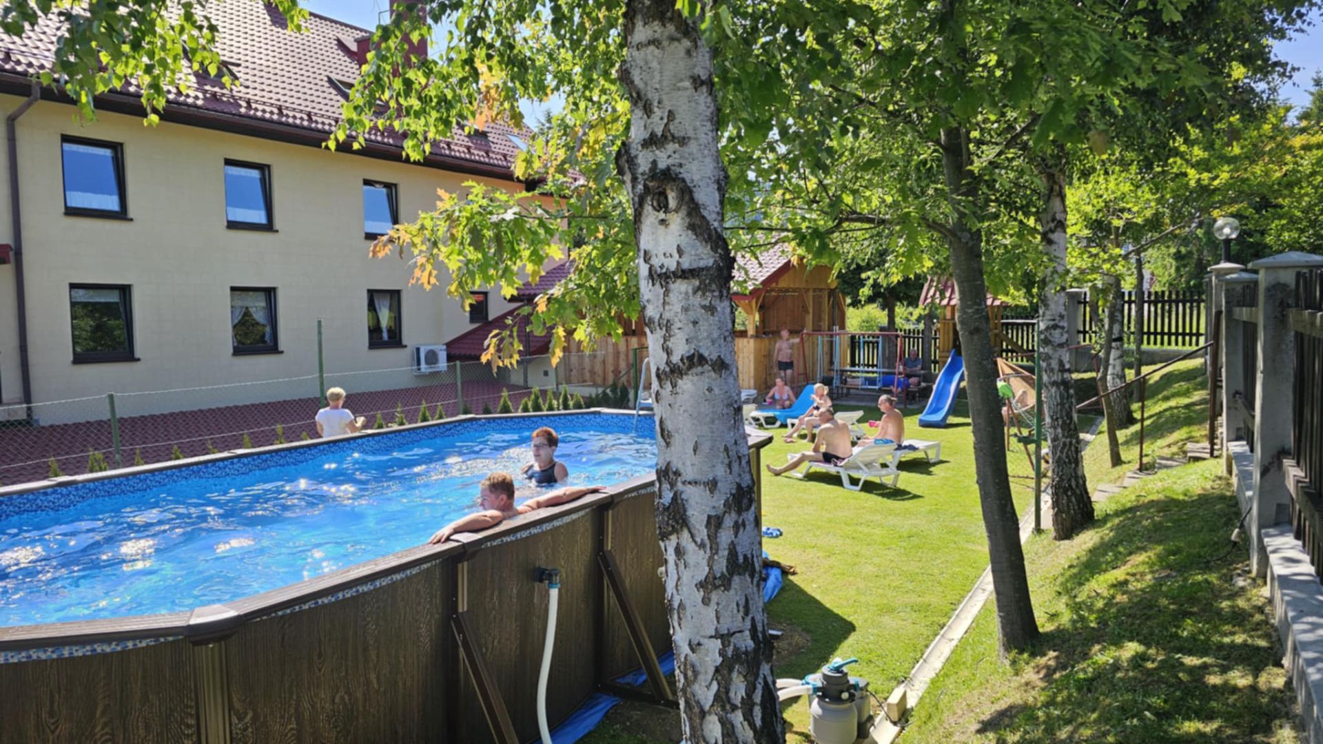 Odkryty basen w ogrodzie Willi OdNova w Tyliczu — goście w wodzie i na leżakach, w tle plac zabaw ze zjeżdżalnią